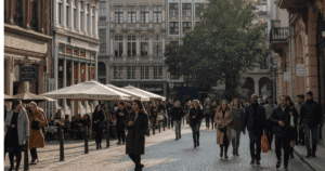 مشهد حضري في بروكسل، يظهر مباني تاريخية وطرقات نظيفة، مع أشخاص متنوعين من جنسيات مختلفة يمشون ويستمتعون بالمدينة. في الخلفية، تظهر مقاهي ومطاعم، وأشجار خضراء، مع لمسة ضوء طبيعي يعكس أجواء يوم معتدل في بلجيكا. المشهد يعكس الثقافة المتعددة اللغات، الحياة الاجتماعية النشطة، والتنوع في البلاد.