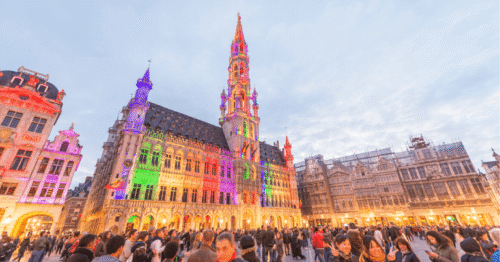 "صورة بانورامية ملونة لعشرة معالم سياحية شهيرة في بلجيكا، مع التركيز على اتوميوم في بروكسل في المقدمة، وخلفه الجراند بلاس وكنائس غينت وقنوات بروج. سماء صافية، ضوء طبيعي دافئ، سياح يتجولون ويلتقطون الصور، ألوان زاهية وتفاصيل معمارية دقيقة، أسلوب فوتوريالي واقعي