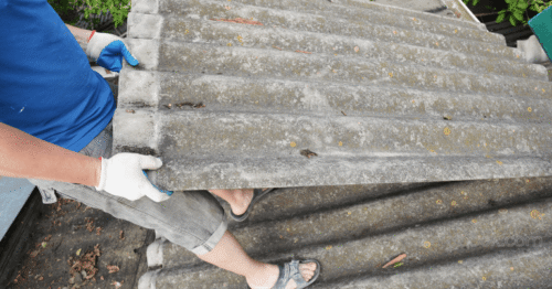 صورة واقعية لمبنى سكني قديم في أوروبا، يظهر سقف من ألواح الأسبستوس المتآكلة، مع عامل فحص يرتدي خوذة وقفازات وقناع واقٍ، يحمل لوحة فحص أو جهاز تقييم، في مشهد نهاري هادئ. أجواء جدية، ألوان محايدة، أسلوب تصوير صحفي واقعي، بدون نص أو شعارات، جودة عالية، مناسبة لمقال إخباري عن الصحة والسلامة في السكن.