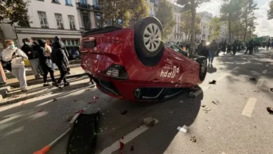 سيارة حمراء مقلوبة على سقفها وسط شارع في بروكسل مع انتشار الحطام ووجود أشخاص في المكان