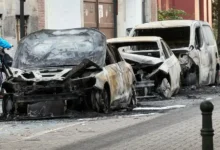 سيارات محترقة ومتضررة بالكامل متوقفة على جانب شارع مع آثار الحريق والحطام حولها